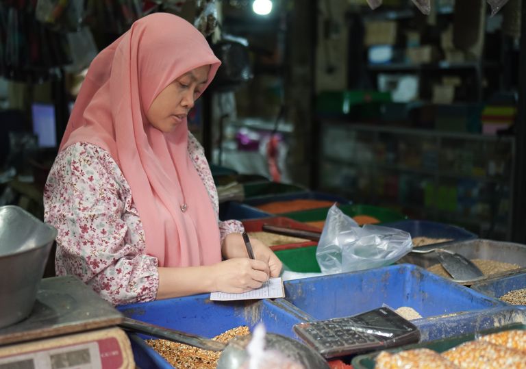 A woman in a hijab calculates sales in an Indonesian market. Vibrant and focused.