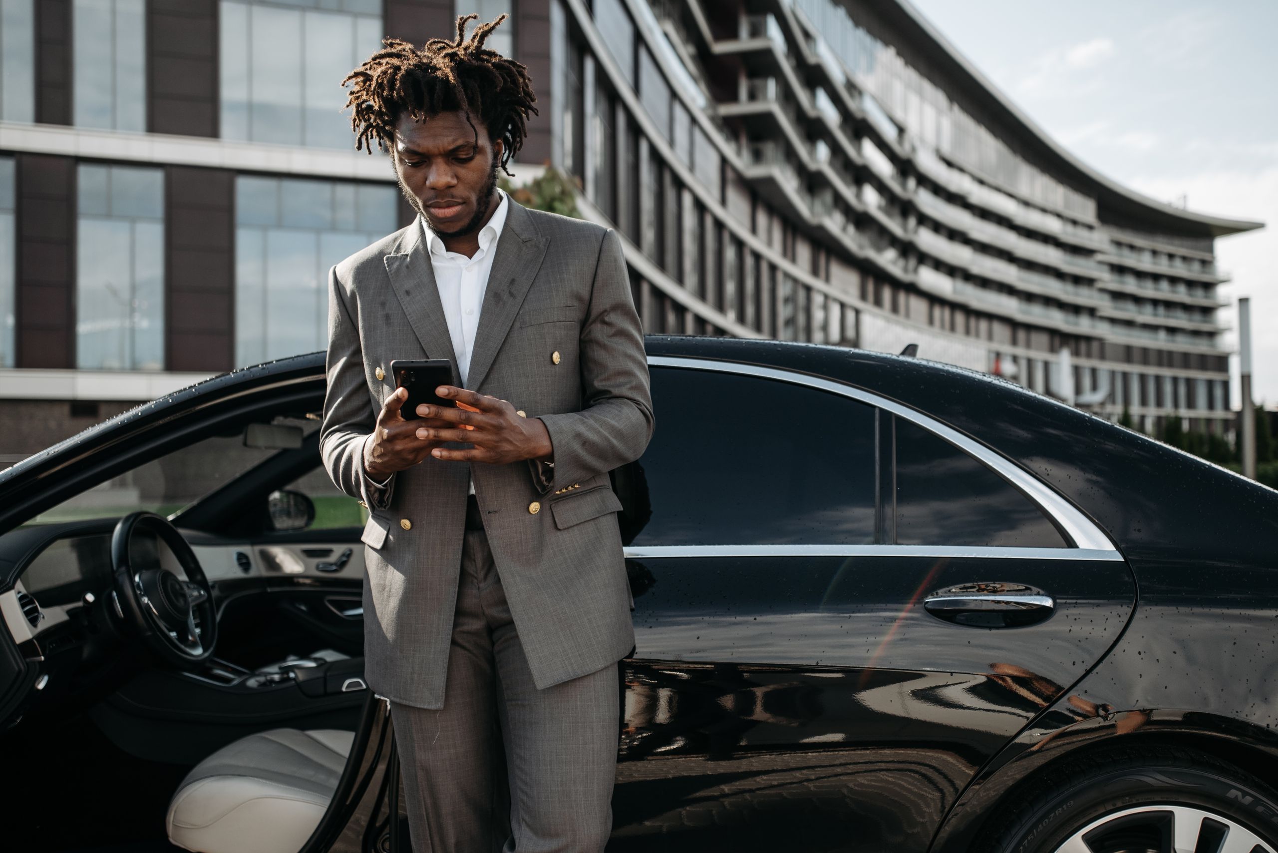 Professional man in a gray suit using his phone next to a luxury car.
